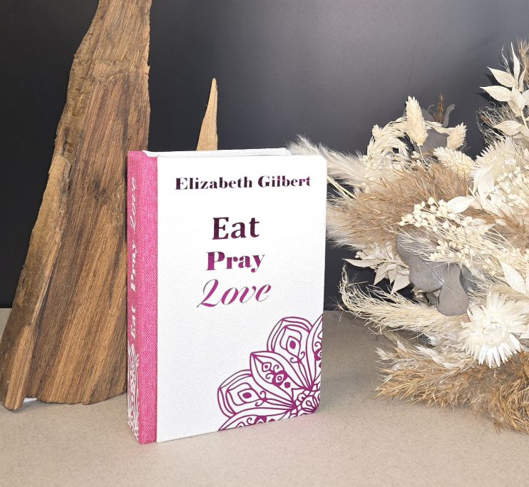 Buch "Eat Pray Love" von Elizabeth Gilbert neben einer dekorativen Pflanze und Holzskulptur.