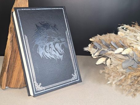 The Witcher - Pentalogie - Andrezj Sapkowski Schwarzes Buch mit einem Wolf-Motiv, dekorativ neben Naturmaterialien platziert.