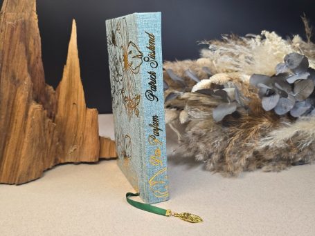 Das Parfum - Patrick Süskind Blaugrünes Buch mit goldener Prägung und grüner Lesezeichen, im Hintergrund braune Holzstücke.