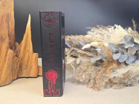 Schwarzes Buch mit rotem Titel und ornamentalem Design, neben einer Holzskulptur.