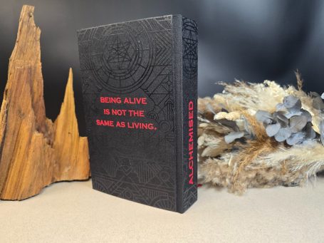 Alchemised - SenLinYu Schwarzes Buch mit rotem Text "Being Alive is not the same as Living" und Naturdekoration.