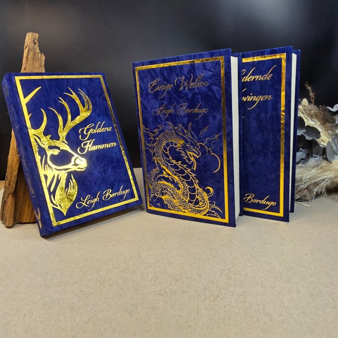 Drei elegante, blaue Notizbücher mit goldenen Verzierungen und fantastischen Motiven.