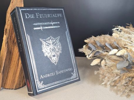 The Witcher - Pentalogie - Andrezj Sapkowski Schwarzes Buch mit Wolfssymbol, umgeben von Holz und Trockenblumen.