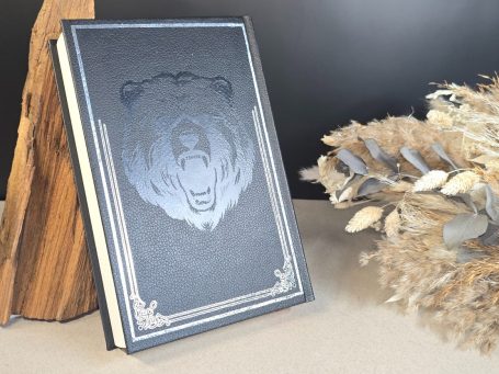 The Witcher - Pentalogie - Andrezj Sapkowski Ein schwarzes Buch mit einem geprägten Bärenkopf und dekorativen Verzierungen.