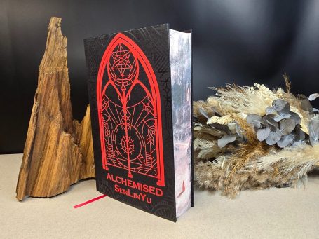 Alchemised - SenLinYu Schwarzes Buch mit rotem, gotischem Design neben einem Holzstück und getrockneten Pflanzen.