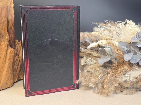 Ein schwarzes Buch mit rotem Rahmen steht neben getrockneten Pflanzen und Holz.